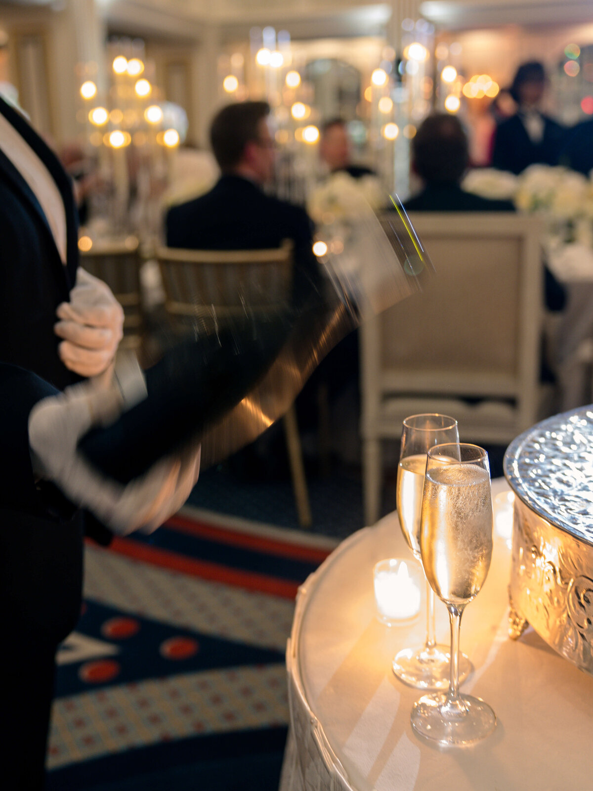 A waiter in a tuxedo pours champagne into glasses on a candlelit table, set in a luxurious restaurant with guests seated at elegantly decorated tables in the background.