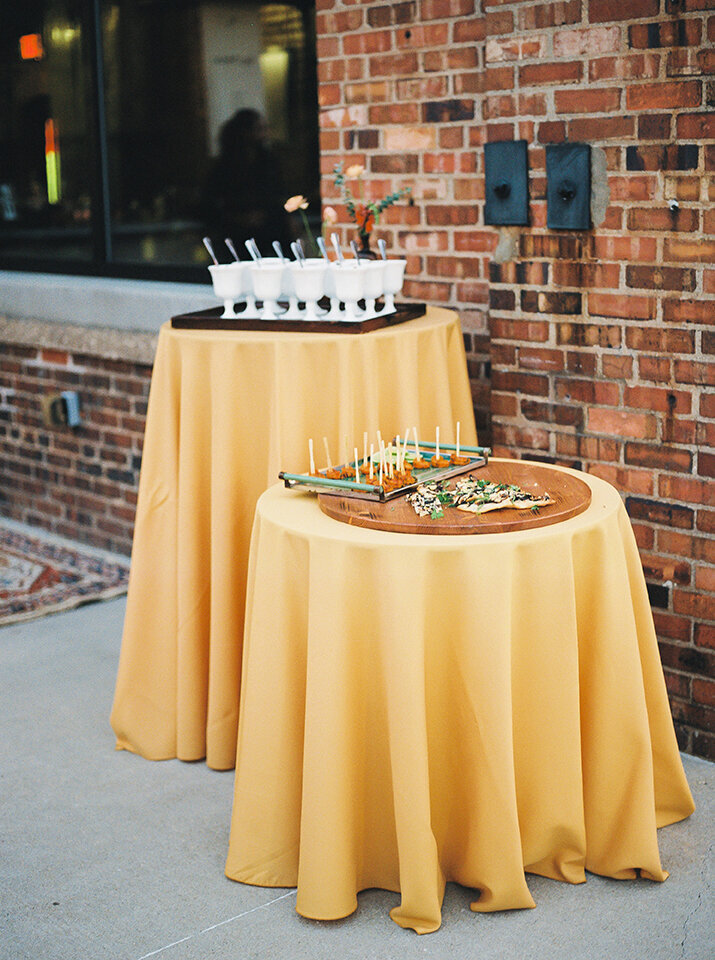 Hors D’oeuvres are set on a wooden tray atop a rounded table with yellow drop clothe.