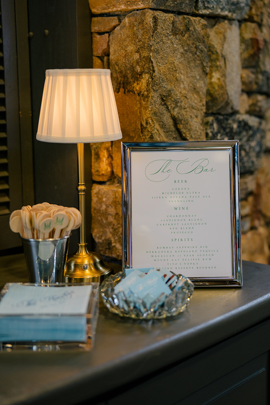  Elegant wedding bar setup with framed drink menu and tabletop lamp against stone wall at Old Edwards Inn wedding reception in Highlands, NC.