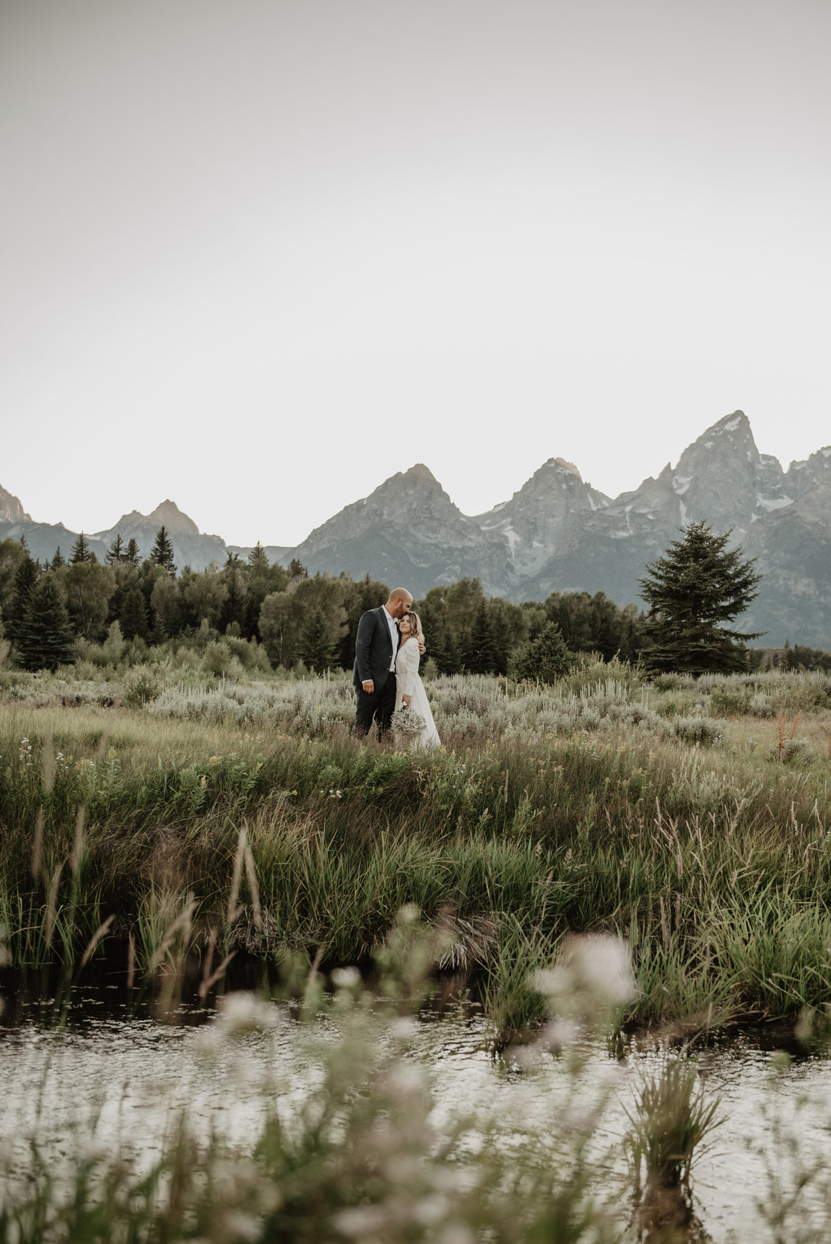 Teton elopement with bride and groom in a field holding each other with the mountains in the background and a creek in the foreground