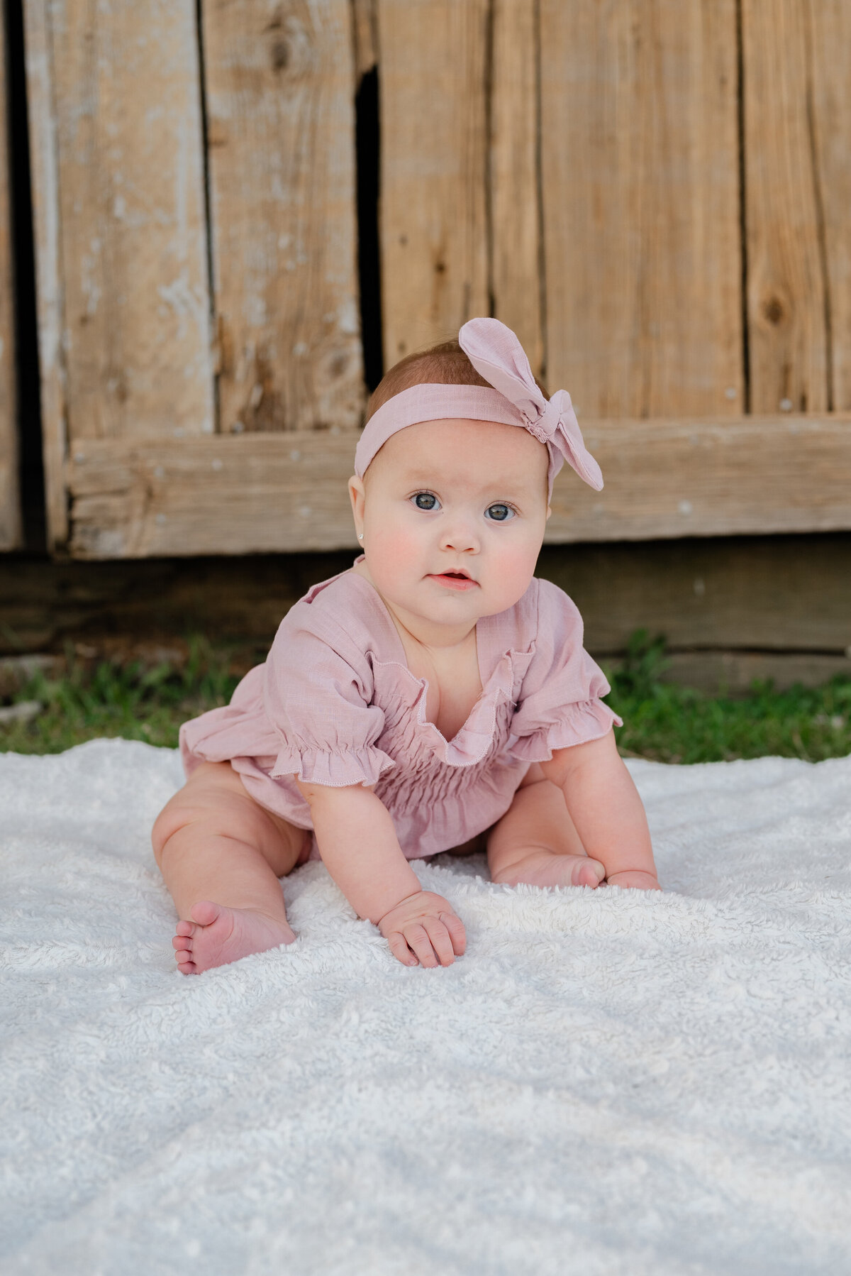 A baby girl wearing blush outfit sitting on a blanket outside.