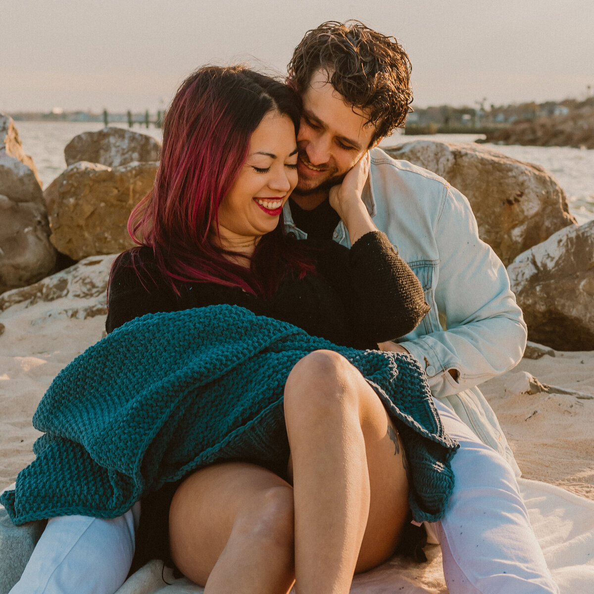 couple cuddling together on the sandy shore of Sylvan Beach in La Porte, Texas, wrapped in a cozy green blanket during their engagement photo session.