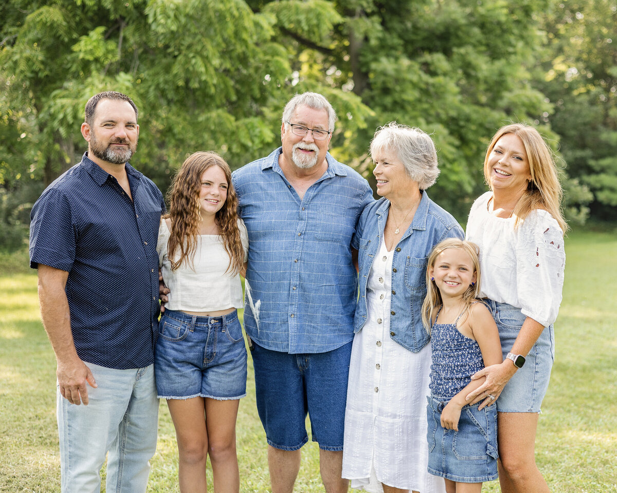 Grandparents, mom, dad, and two daughters standing next to each other with some looking at the camera smiling while others looking at each other.  Wife is wearing a jean skirt, white half sleeve shirt, and has long blonde hair.  Husband is wearing a blue button up short sleeve shirt, blue jeans, and has black gray short hair and beard.  Older daughter is wearing blue jean shorts, a white short sleeve shirt, and has long brown hair.  Grandpa is wearing a light blue and white stripped short sleeve button up shirt, blue jean shorts, and has white short hair and goatee.  Grandma is wearing a white button up dress with a jean jacket over it, with short white hair.  Youngest daughter has a dark blue and white tank top style shirt with a blue jean skirt, and has medium length blonde hair.