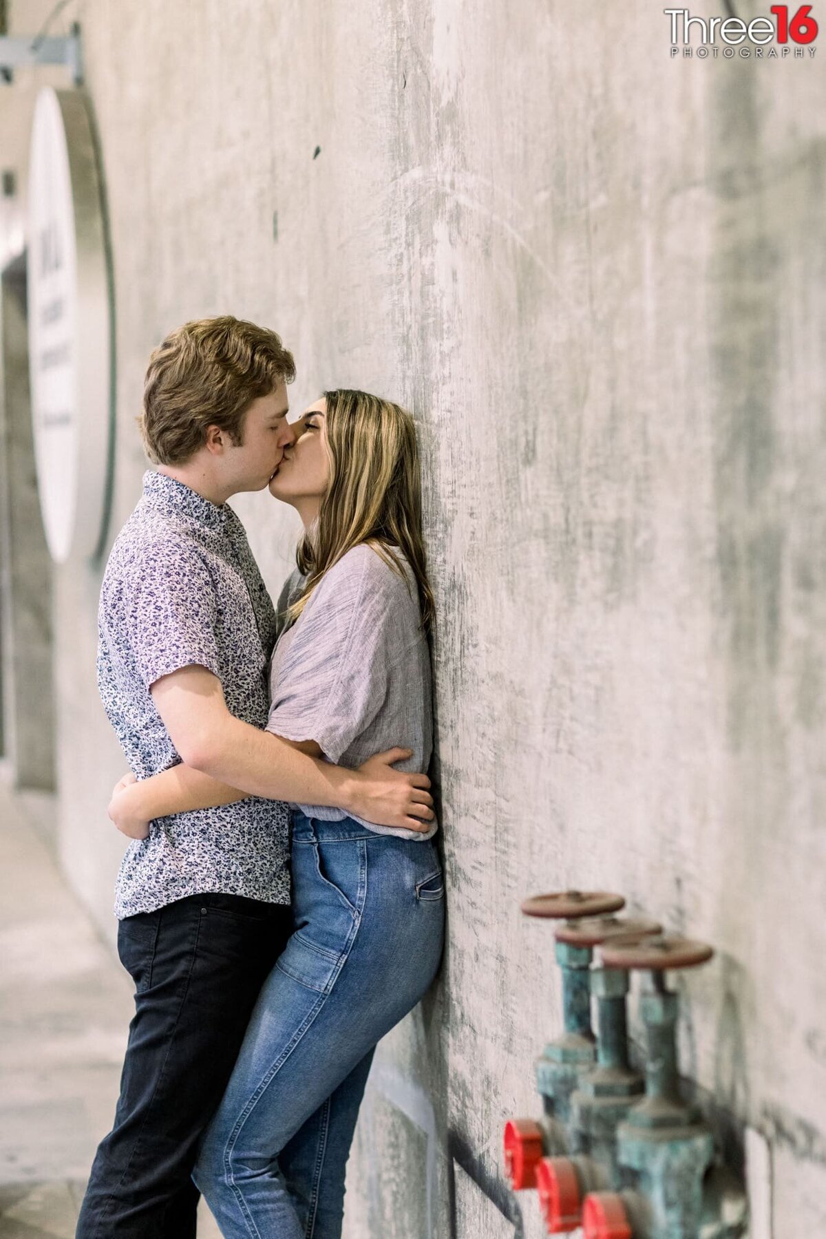 Engaged couple share a romantic kiss as the Bride to be leans back against a cement wall