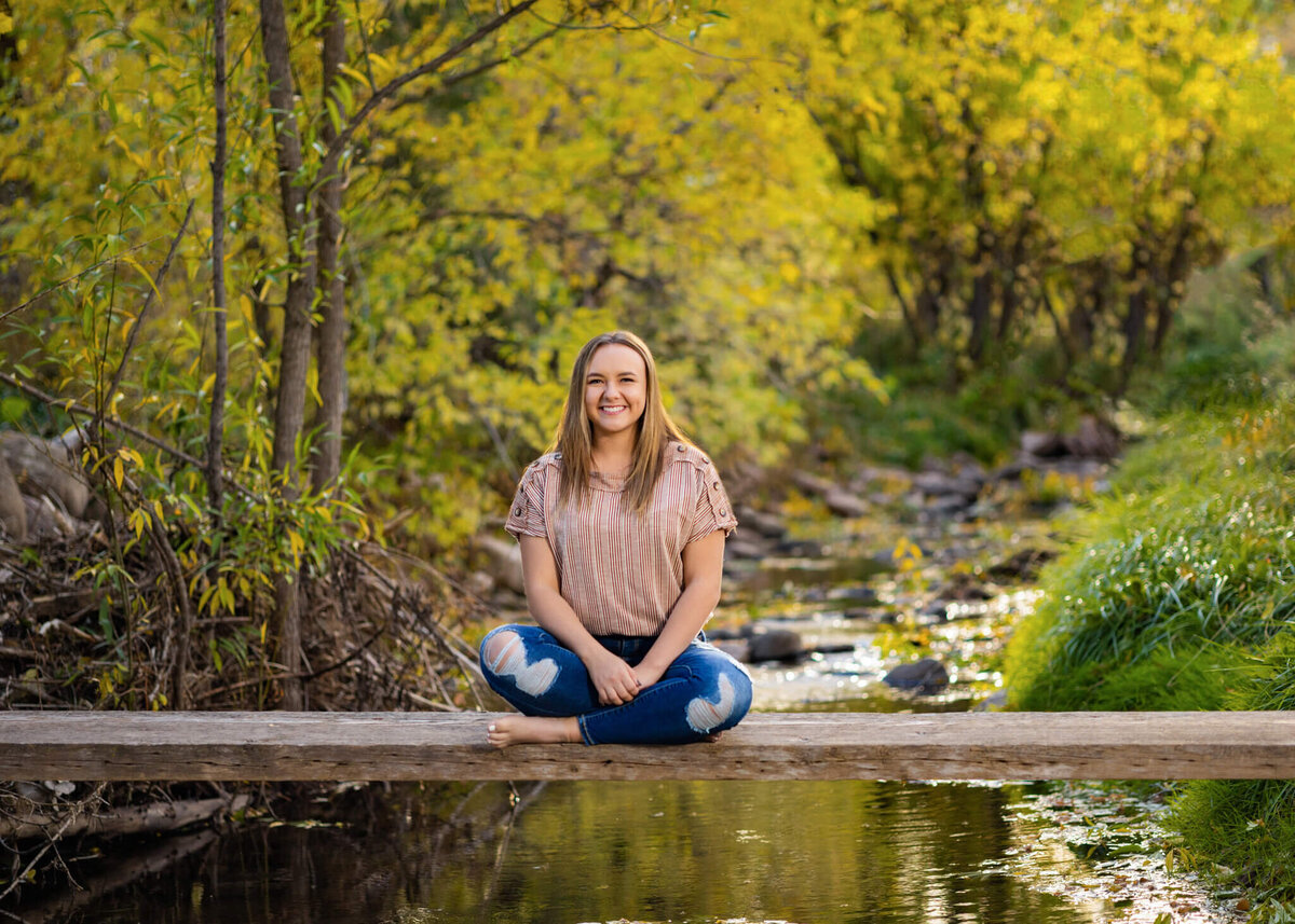 high school senior girl sitting criss cross on a wooden plank over a shallow creek