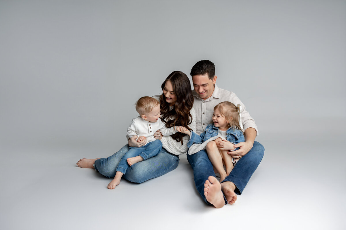 Parents sitting on floor with their two daughters leaning in together, captured in natural studio light. Photographed by Yucaipa family photographer Kaitlyn Dawn Photography.