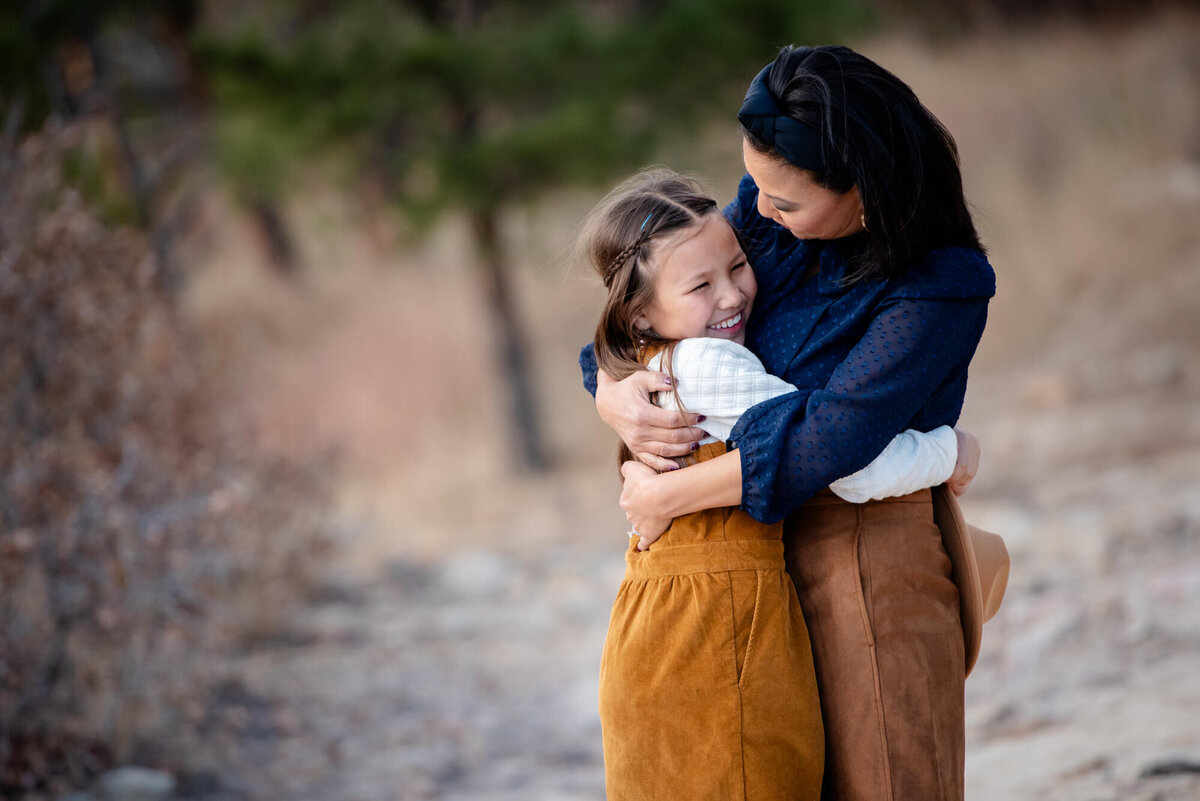 A mother in a blue top hugs her daughter in an orange dress while they laugh together