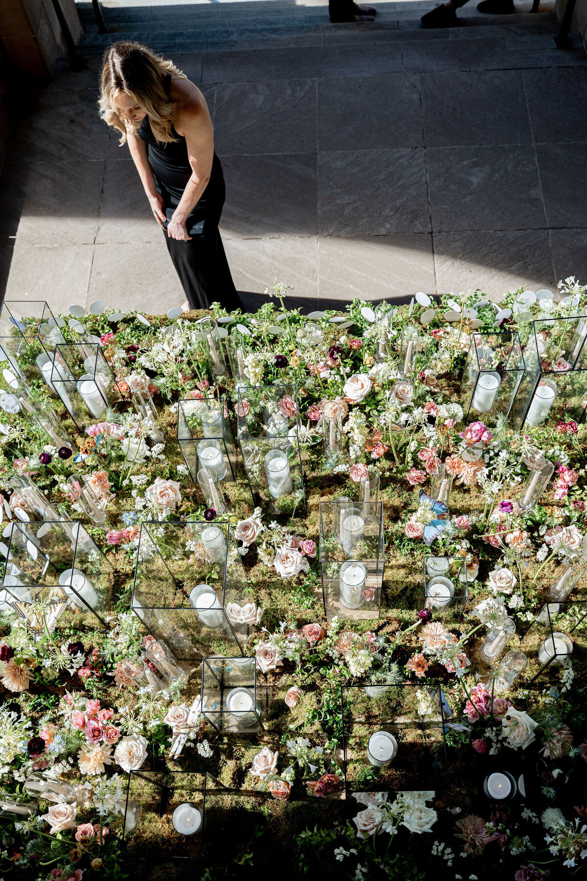 This photo was taken at Washington National Cathedral in Washington D.C. by photographer Twah Dougherty. A woman in a black dress stands on a stone floor, looking at a decorative arrangement consisting of numerous glass vases filled with white and pink flowers, candles, and greenery. The display is set in a pattern and bathed in natural sunlight. Additionally, Current Films also worked as videographers at this event.