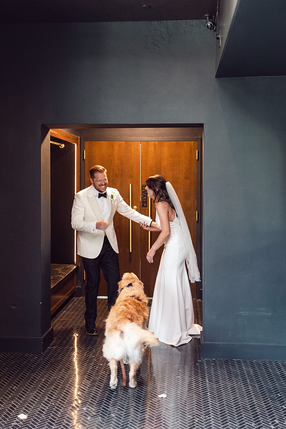 The bride, wearing a silk halter top sheath wedding dress and long veil and the groom wearing a tuxedo with a white jacket share a first look with their best man, their golden retriever Frank in the entry way of Clementine Hall.