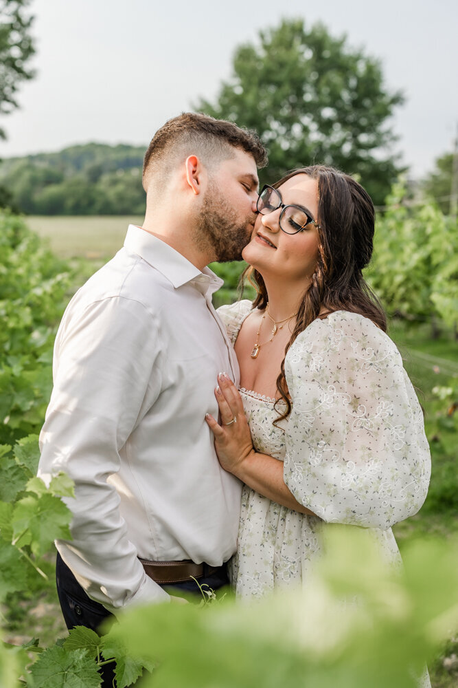 Man kissing his fiance on the cheek in a vineyard
