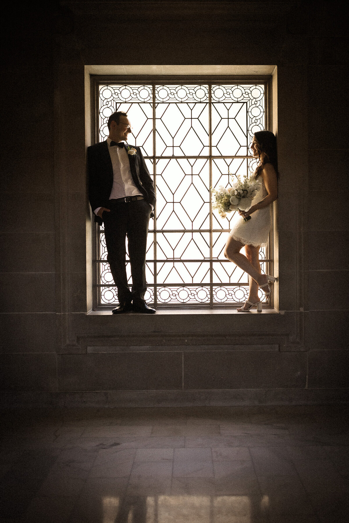 Couple stands in window at their San Francisco City Hall Wedding. Bride is in short white dress and groom is in a black suit.