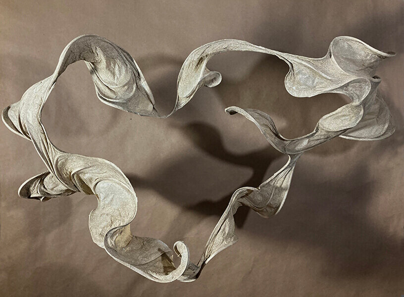Circular, wave-like sculpture formed from handmade abaca paper, photographed from above against a brown background.
