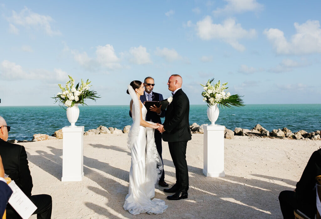 The bride and groom stand hand in hand as they exchange vows with the ocean waves behind them, creating an unforgettable Florida Keys wedding moment. Destination wedding photography by Claudia Amalia Photography.