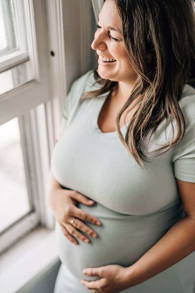 A radiant expecting mother laughs while she looks at the window during a session with Minneapolis maternity photographer Kate Simpson.