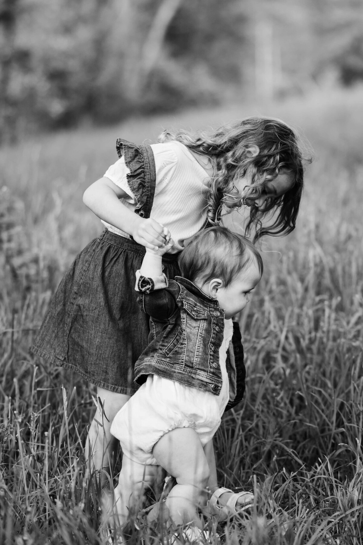 A young girl assists a toddler with walking in a grassy field, both are dressed in denim attire. This moment captured by a skilled Pittsburgh, PA photographer showcases the beauty of family connections.