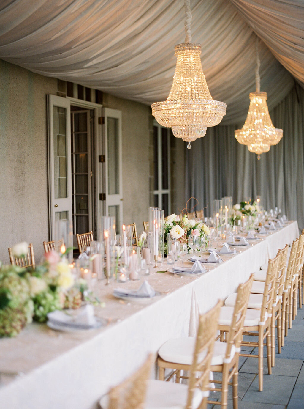 Wedding Photographer RI | Elegant table setting with white tablecloth, crystal chandeliers, and gold chairs. Decor includes white flowers, greenery, and candles.