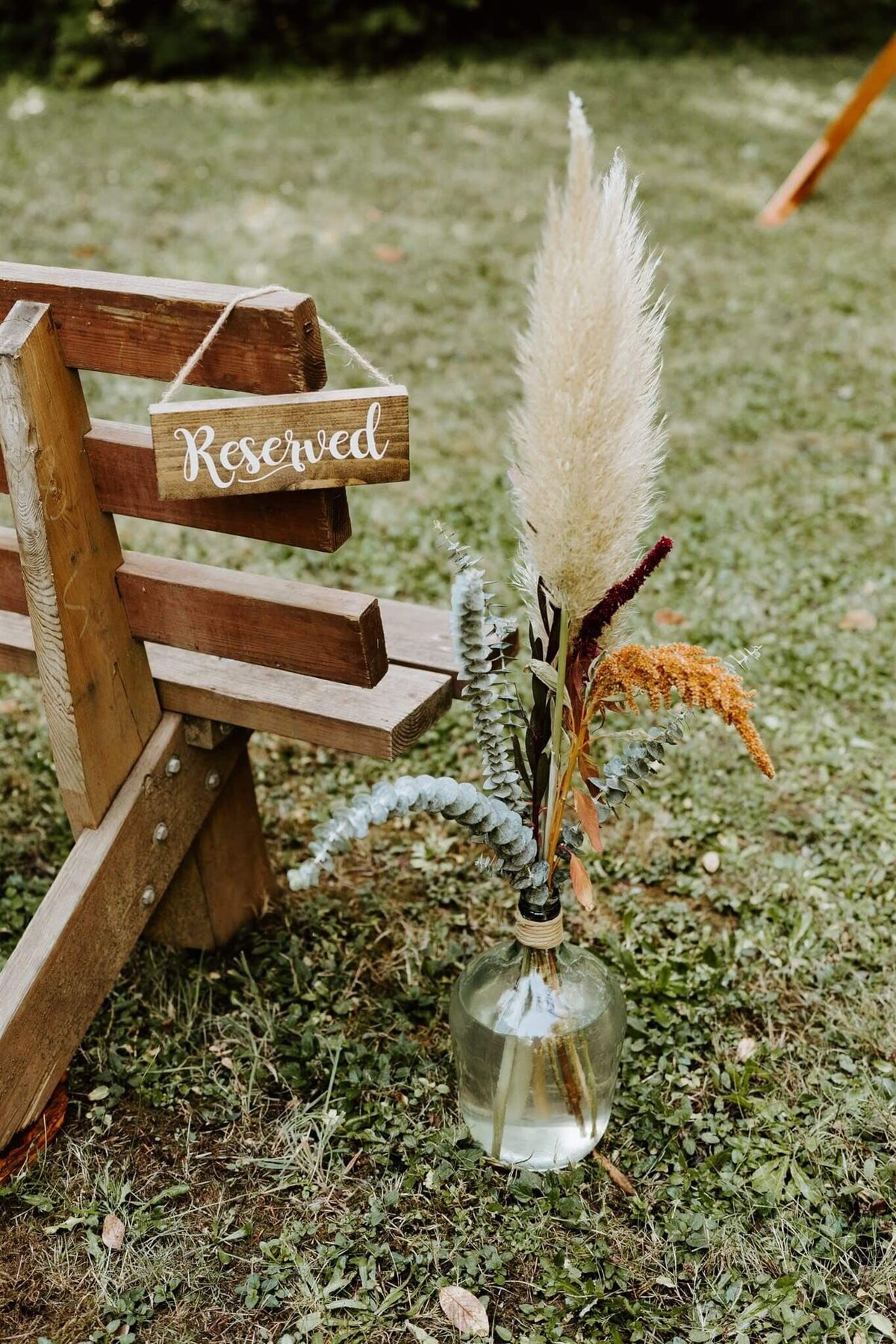 Boho wedding floral design with a bud vase with pampas grass, eucalyptus and more  sit by a reserved seat at an outdoor wedding ceremony.