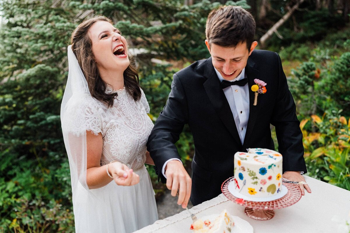 couple in wedding attire laughing while cutting the cake in Mount Rainier National Park