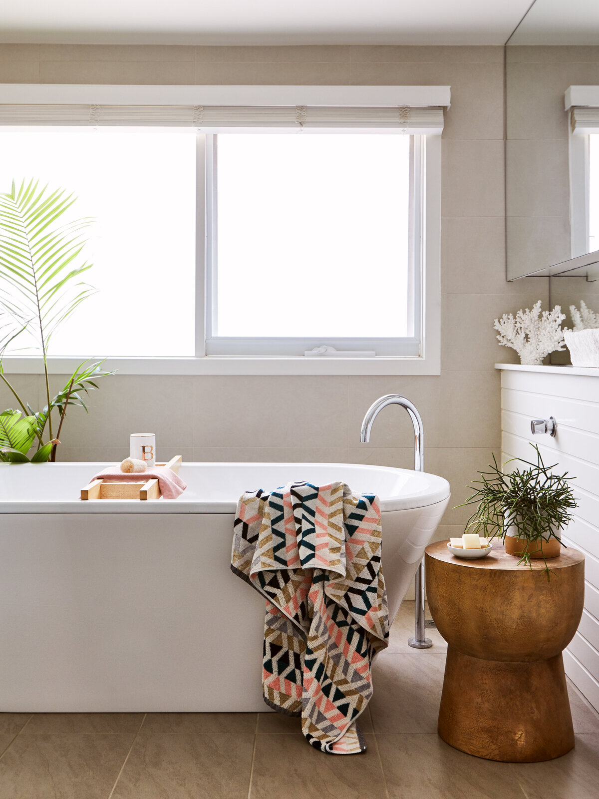 Freestanding bath styled with patterned towel, timber stool and greenery in a light-filled Eynesbury bathroom.