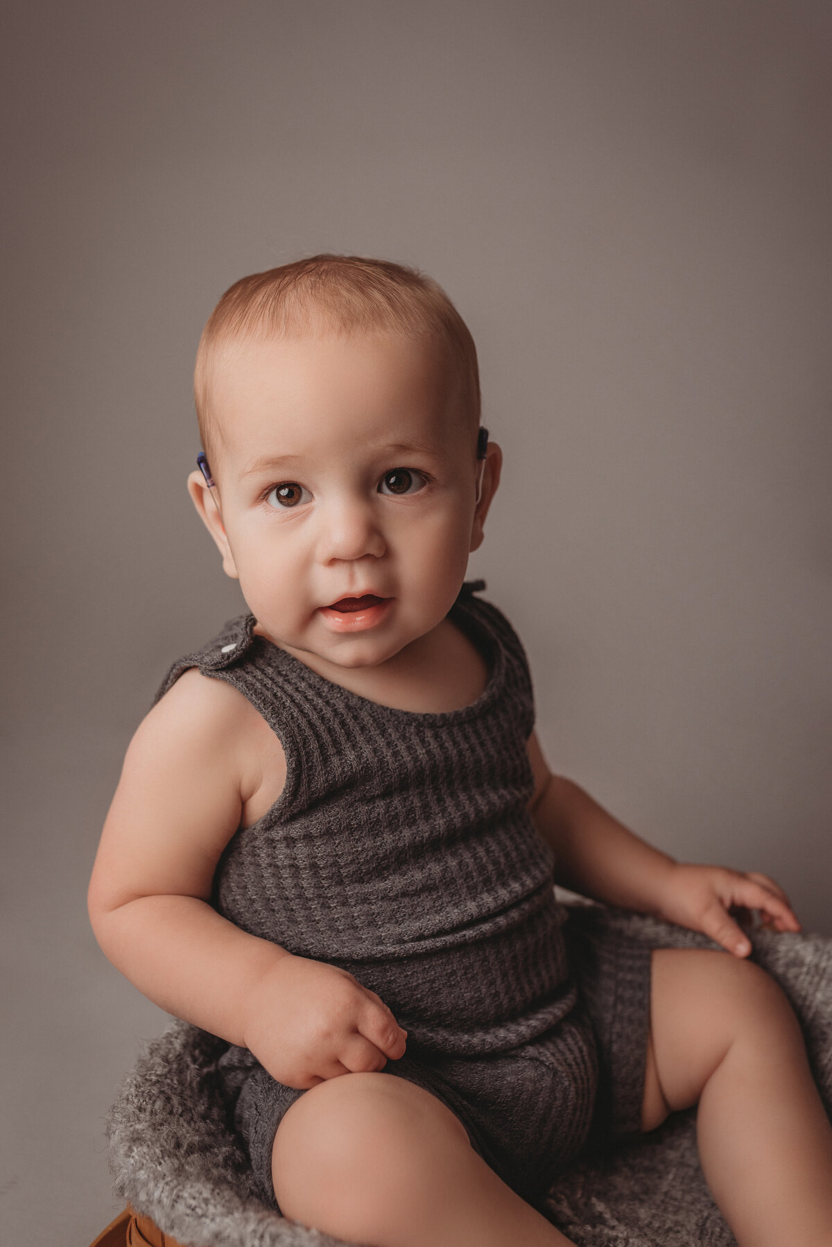One year old baby boy wearing gray waffle knit romper sitting in basket posing for his portraits on a light gray backdrop
