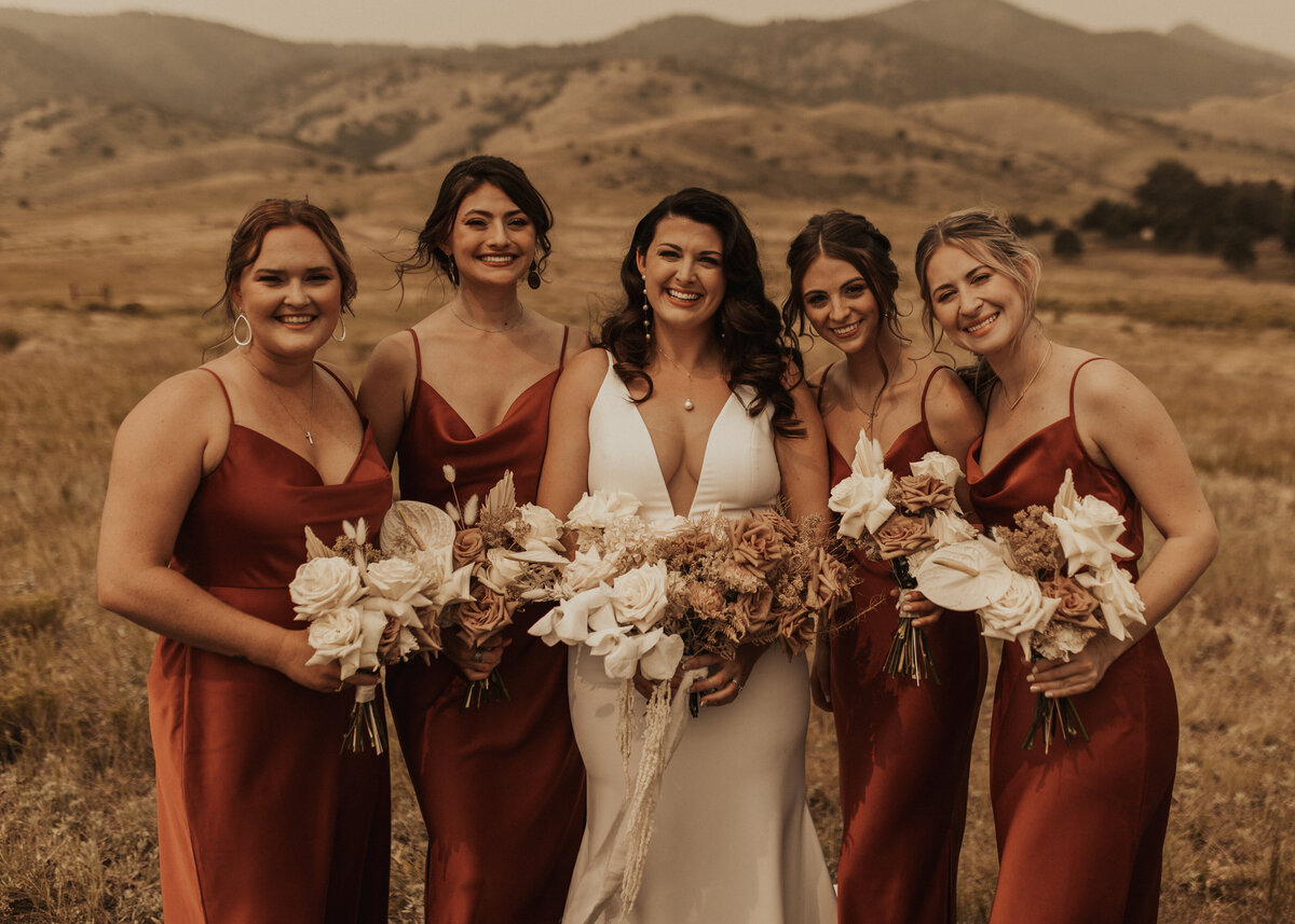 Bride and bridesmaids stand in a field  holding flowers smiling