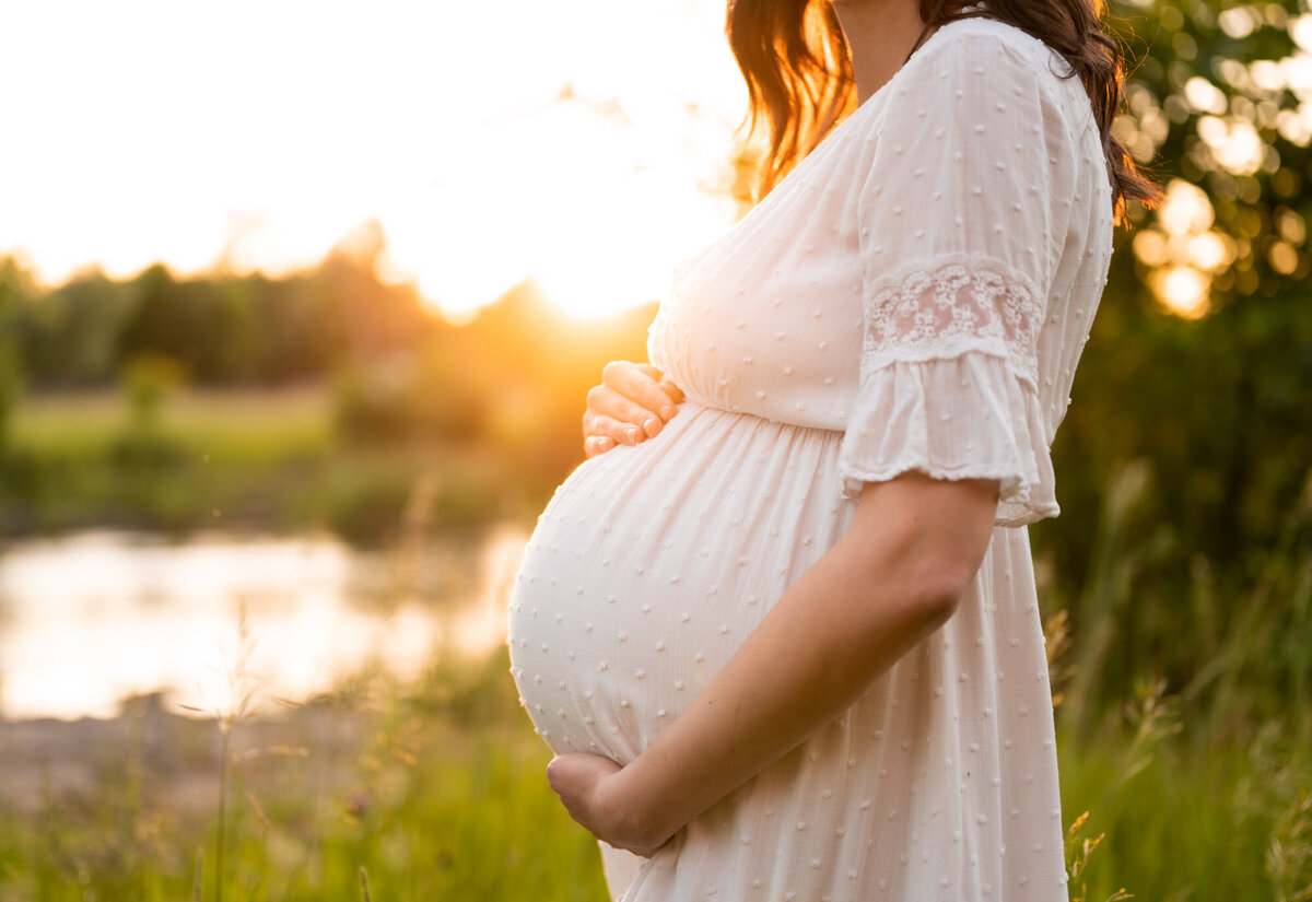 Pregnant belly in a white dress lit up by golden hour sun.