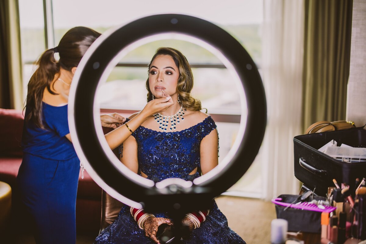 Indian bride getting her makeup done by a makeup artist in a hotel with a ring light in the foreground 