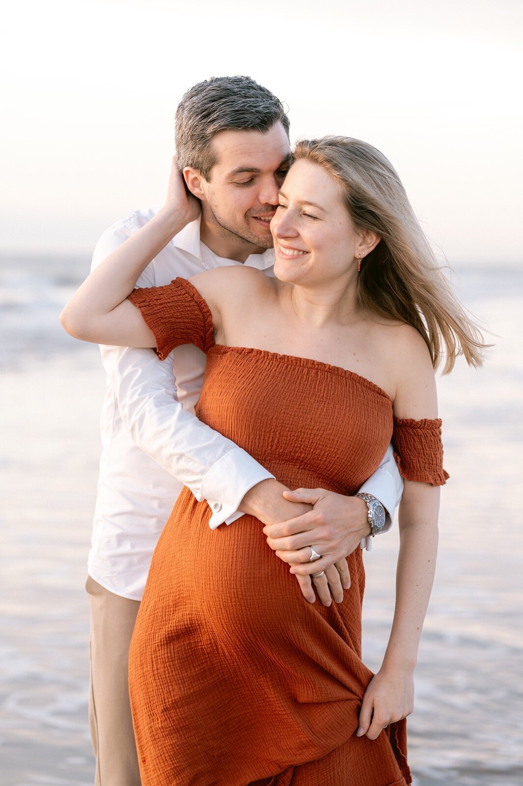Man staat achter zijn zwangere vrouw met zijn armen om haar heen. 