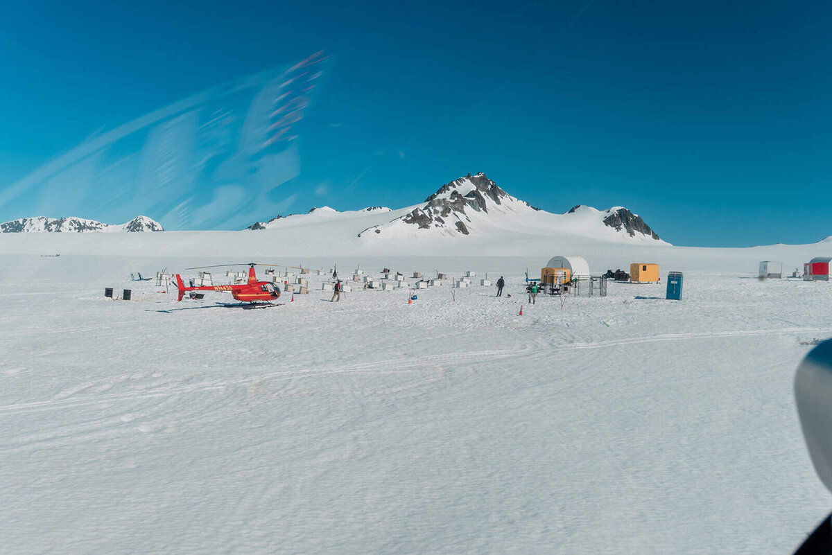 A snowfield glacier stretches as far as the eye can see during a two-day Kenai Fjords luxury elopement.  There is a bright red helicopter near the dogsled camp that is viewable in the distance.  Several dozen small white hutches are visible with a few dogs around. 