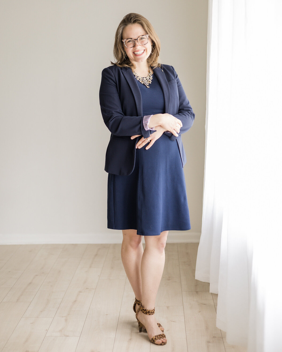 Brunette blonde pregnant female standing with her hands on her stomach, wearing glasses, a dark blue dress with a long sleeve dark blue jacket, smiling at the camera