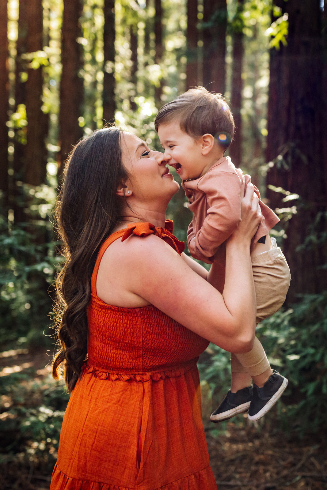 mom in orange dress holding her toddler nose to nose in the woods while they smile at each other