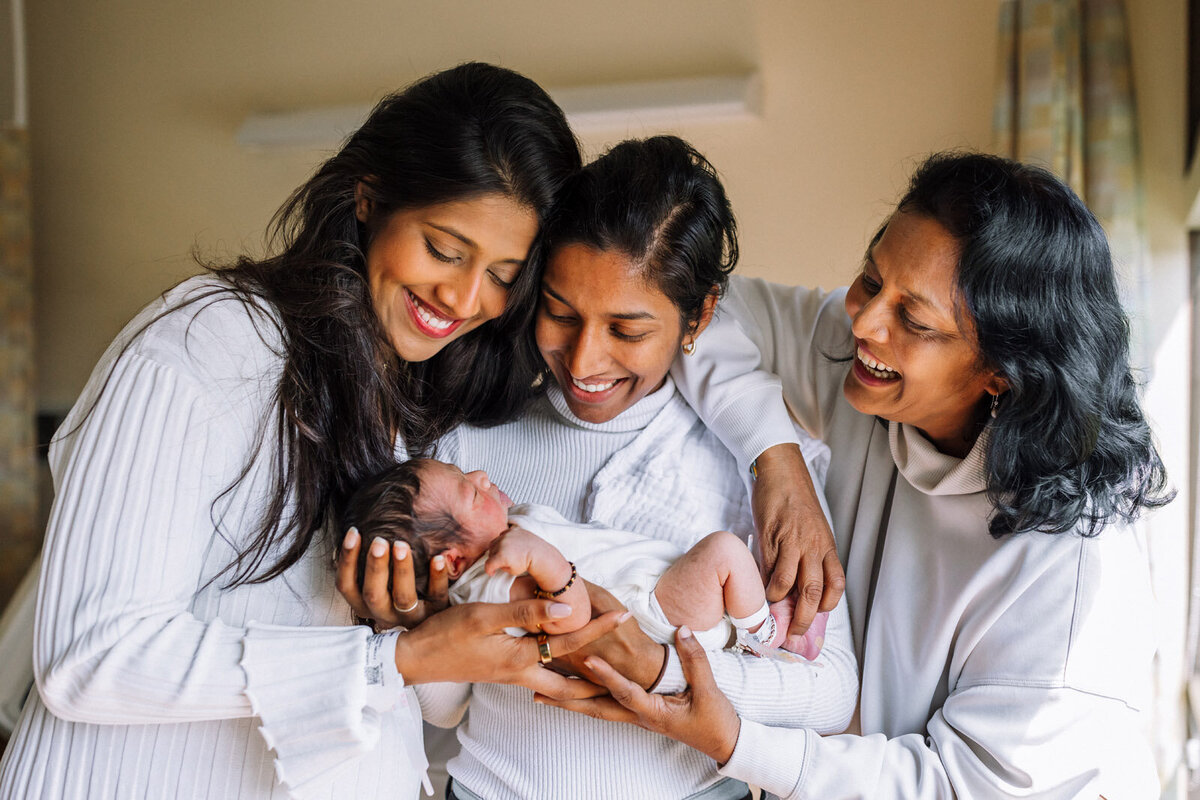 new mom, sister, and grandmother smiling and looking at newborn in hospital