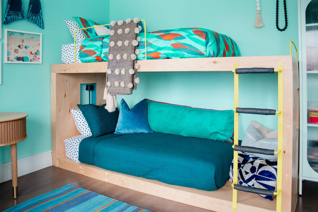 A brightly lit children's bedroom with light turquoise walls features a wooden bunk bed with colorful bedding. The top bunk has a patterned comforter with teal, orange, and white fish-like shapes, along with patterned pillows and a textured gray throw with large pom-poms. The bottom bunk has a dark teal quilted comforter and several pillows in shades of teal, dark blue, and white with black polka dots and geometric patterns. A yellow ladder provides access to the top bunk. To the right of the bunk bed, a light mint green cabinet with arched glass doors stands against the wall. A long beaded necklace hangs on the wall near the cabinet. A glimpse of a wooden floor is visible.