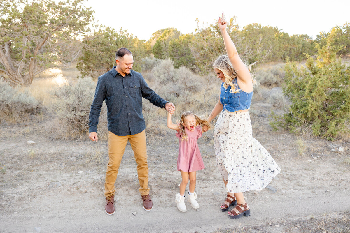 A father and mother hold their daughter’s hands as she joyfully swings between them, laughing in the middle of a dirt path.