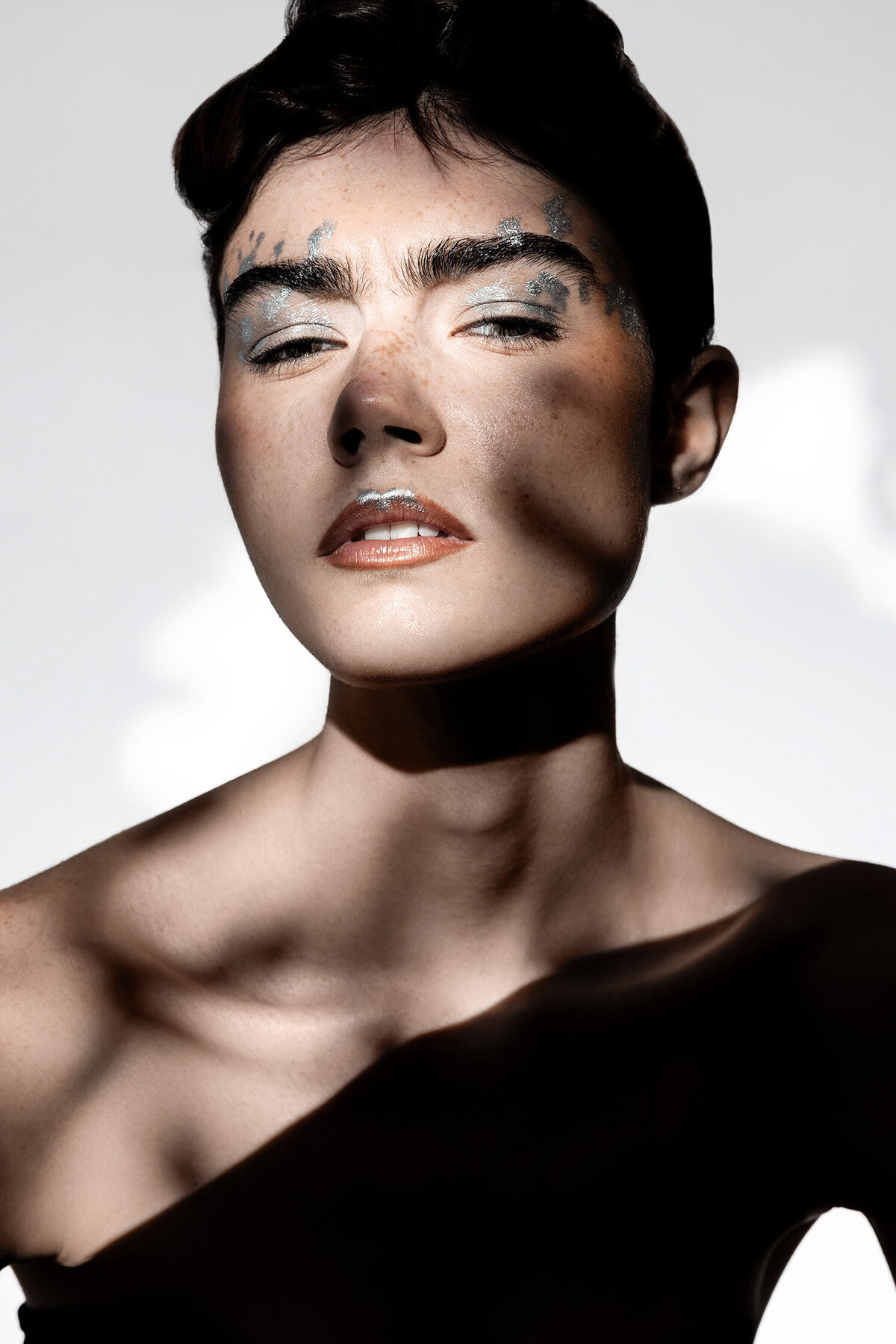 Beauty photography portrait with freckled model wearing silver metallic makeup under dramatic shadows