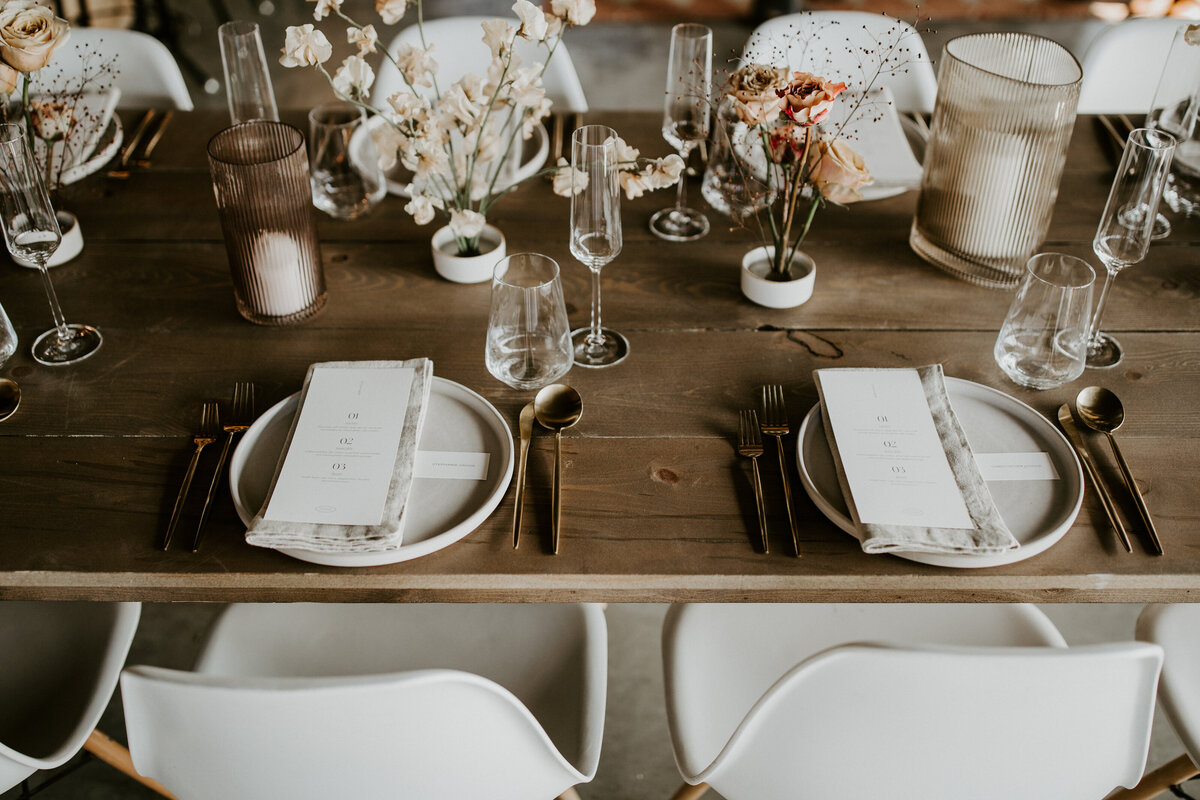 White dinner place cards set on linen napkin and white plates on a wooden table with white flowers and white chairs.