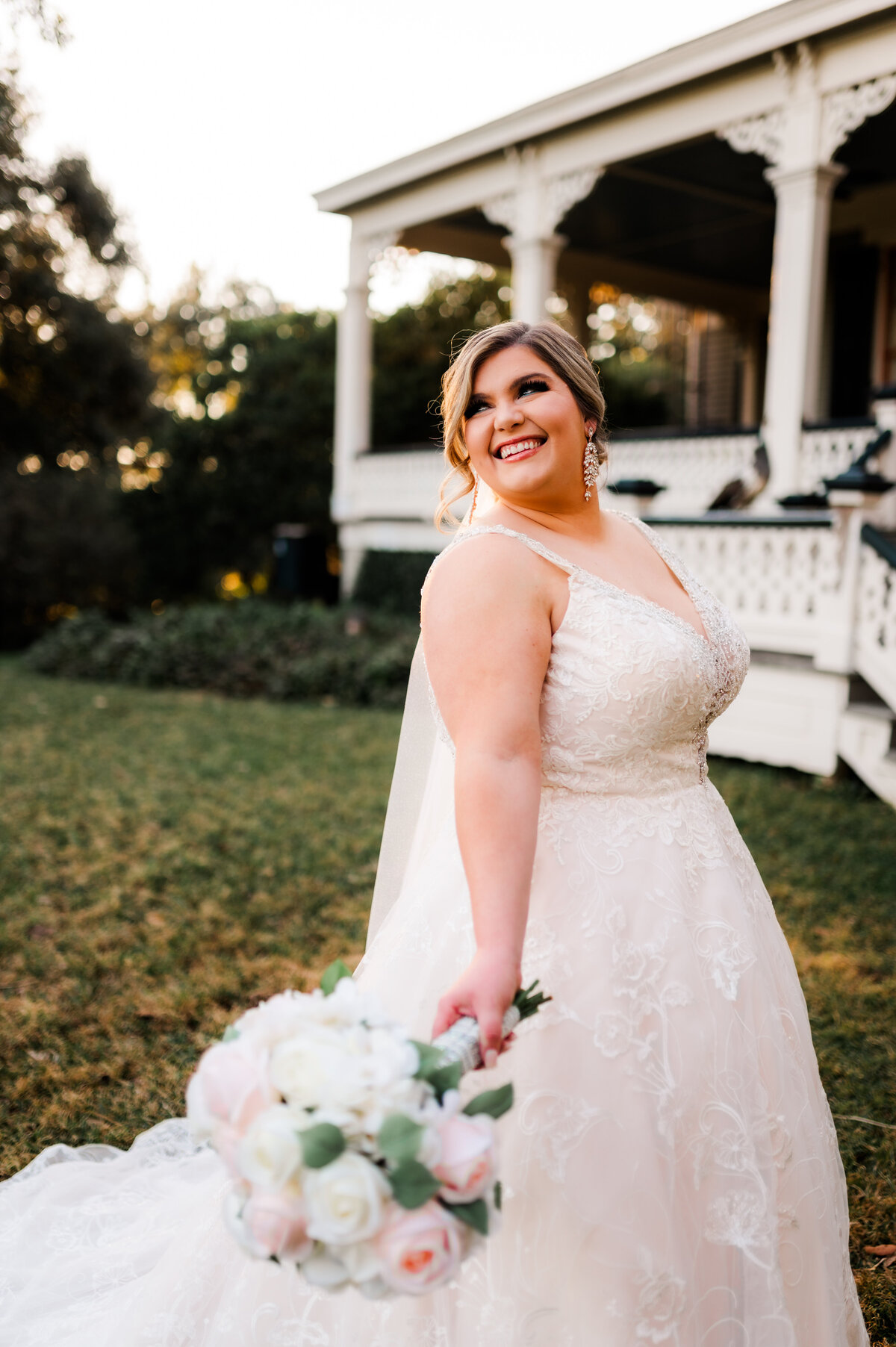 Little Rock wedding photographer captures bride standing outside of her wedding venue on the lawn while the sun sets through the trees in the distance