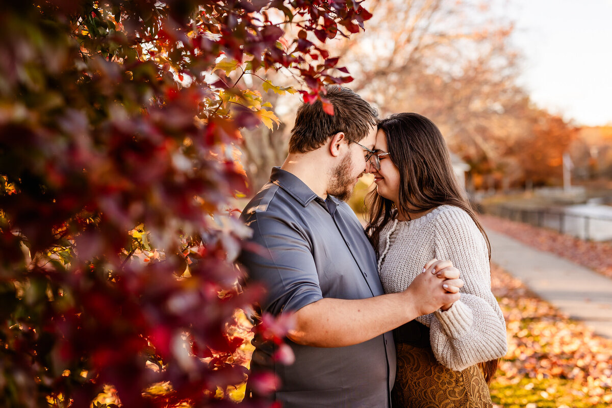 Women leaning on fiancee behind beautiful fall tree with vibrant red call color at sunset engagement session