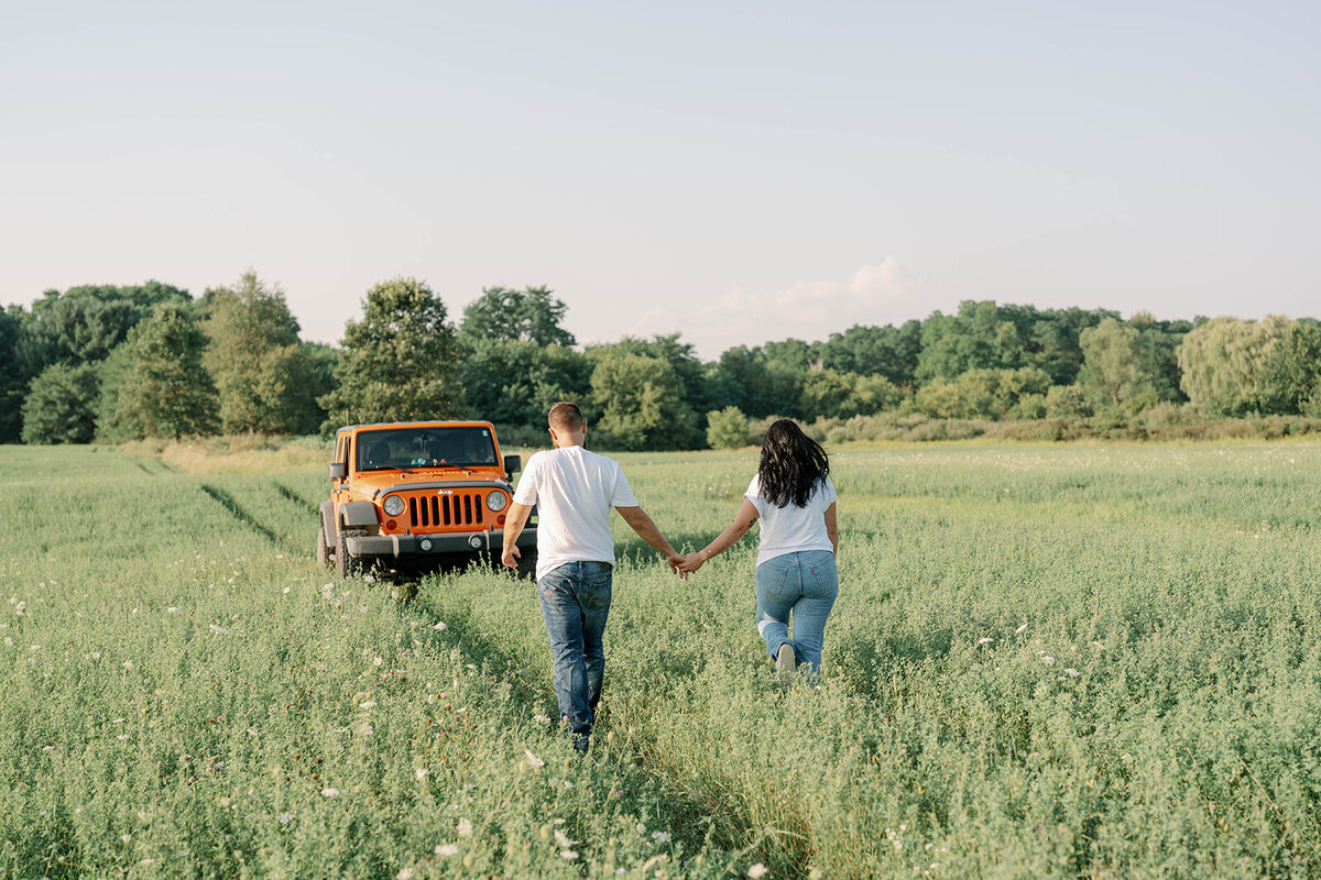 Kali and Joe walking toward a vintage orange jeep parked in an open field during their Detroit engagement session.