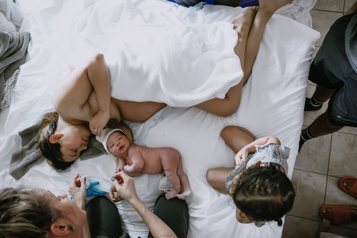Mother and child lay on the bed with eldest child and midwife nearby