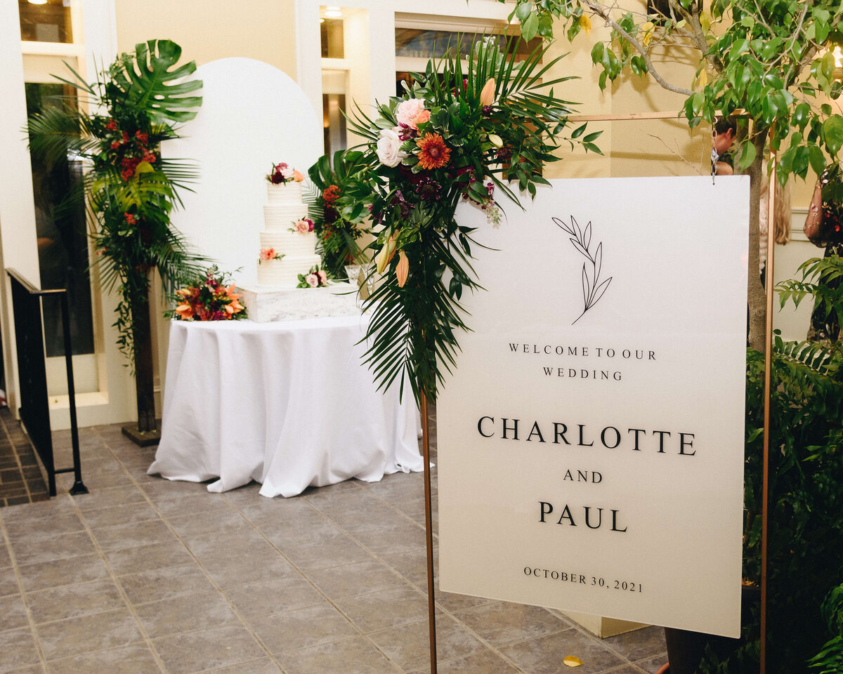 A wedding welcome sign with Charlotte and Paul stands by a floral arrangement. In the background, a white table displays a tiered wedding cake decorated with flowers.