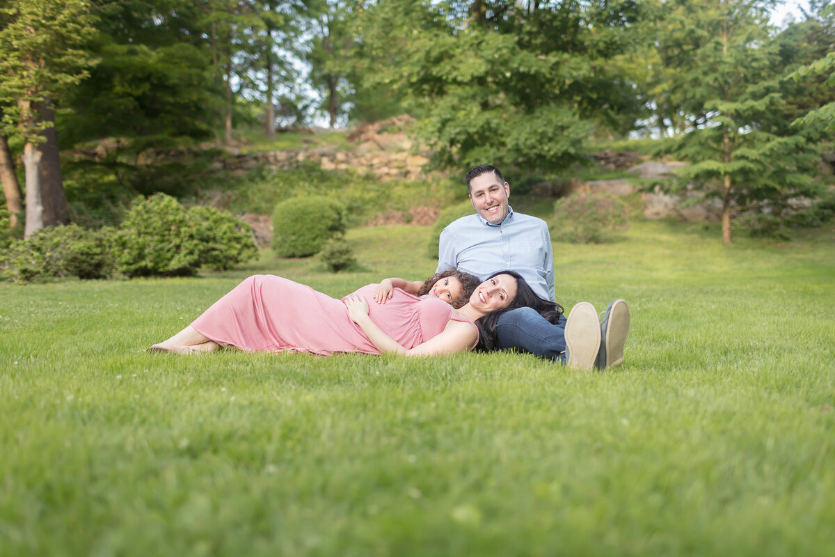 This family maternity portrait was photographed in a beautiful, lush green park. The father sits in the grass, leaning back on his hands, as his wife and daughter lay on his lap. The mother to be is wearing a peachy pink, babydoll style maxi dress. She and her curly-haired daughter are smiling at the camera, each with a hand on Mom's belly.