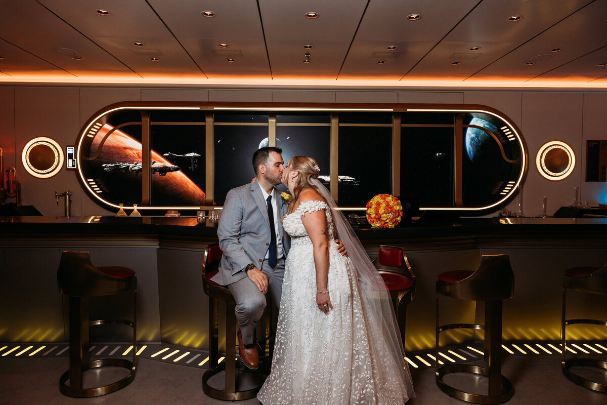 Bride and groom share a kiss in the Hyperspace Lounge aboard the Disney Wish cruise ship with space-themed backdrop.