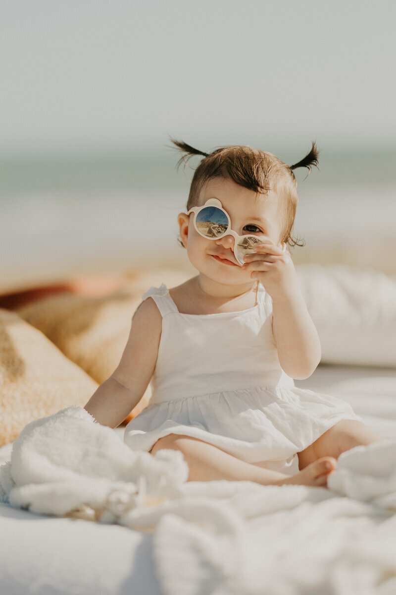 Petite fille en robe blanche avec deux couette et des lunettes de soleil ronde dans un décor cosy à la plage pour une photo de famille en Vendée.