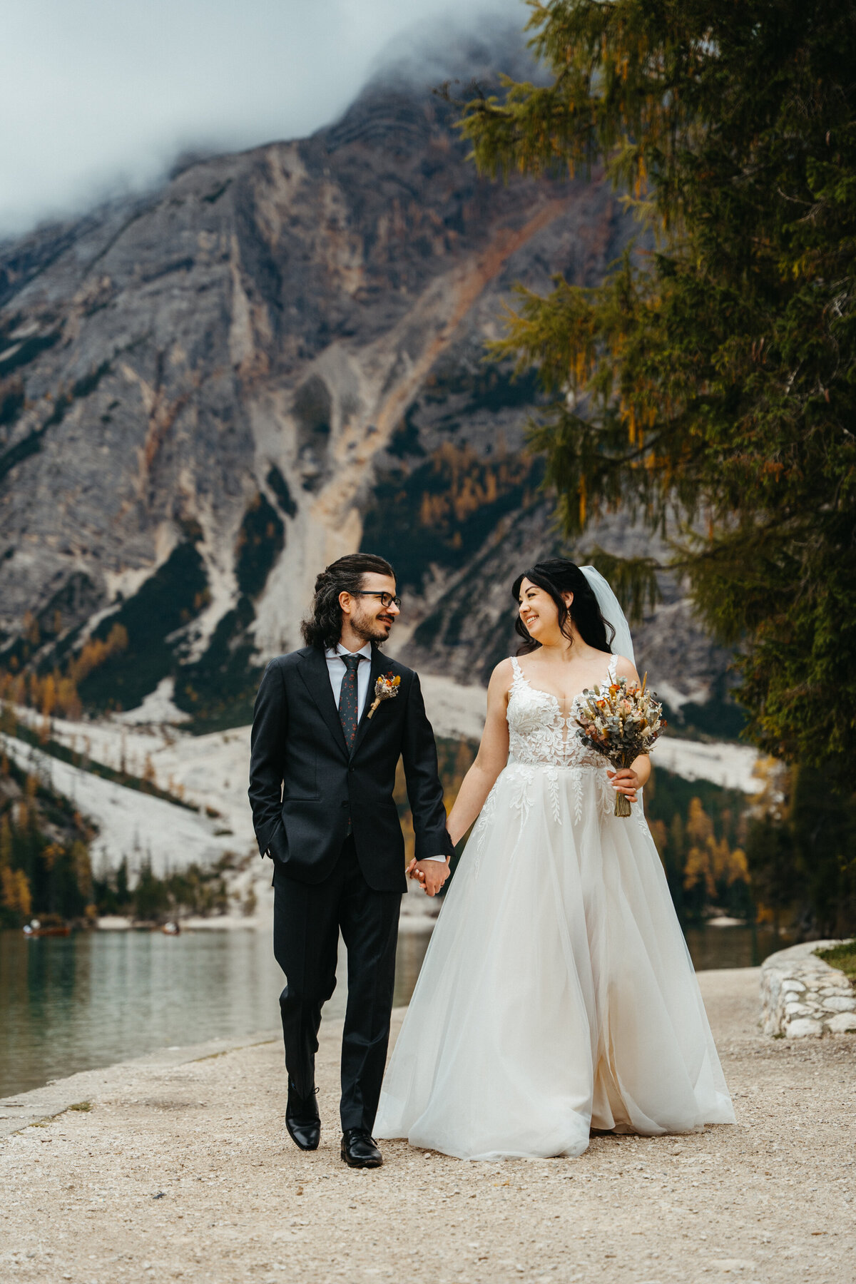 Bride and groom walking through forest beside alpine lake