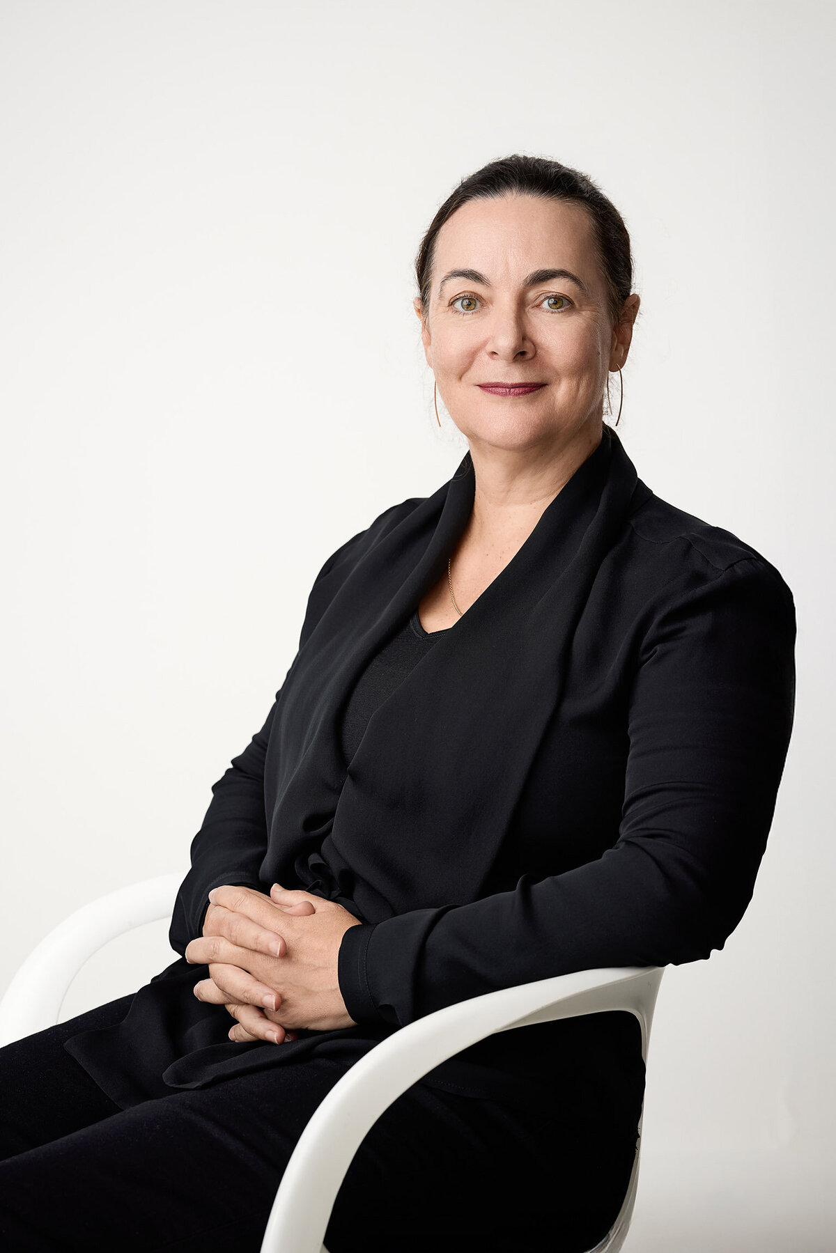 Woman wearing black seated on a white chair against white background. She has a quiet confident gaze looking at camera.