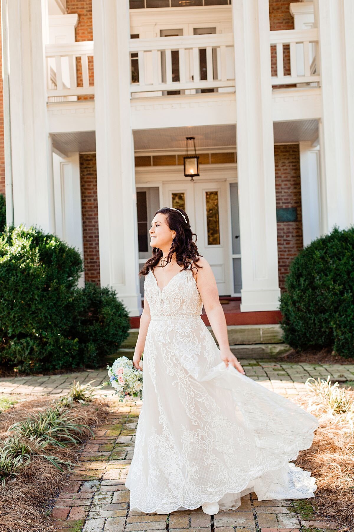 The bride is standing in front of a large white mansion with a two tier porch. She is standing on a stone path with green bushes on either side. The bride is wearing a lace dress with a long flowing skirt that blows in the breeze. The dress has a plunging neckline and spaghetti straps. She is holding a white and blush bouquet down at her side.