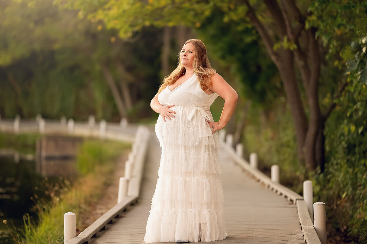Pregnant woman in a tiered white gown standing confidently on the Delafield boardwalk, surrounded by lush green trees and soft sunset light during a serene outdoor maternity session.