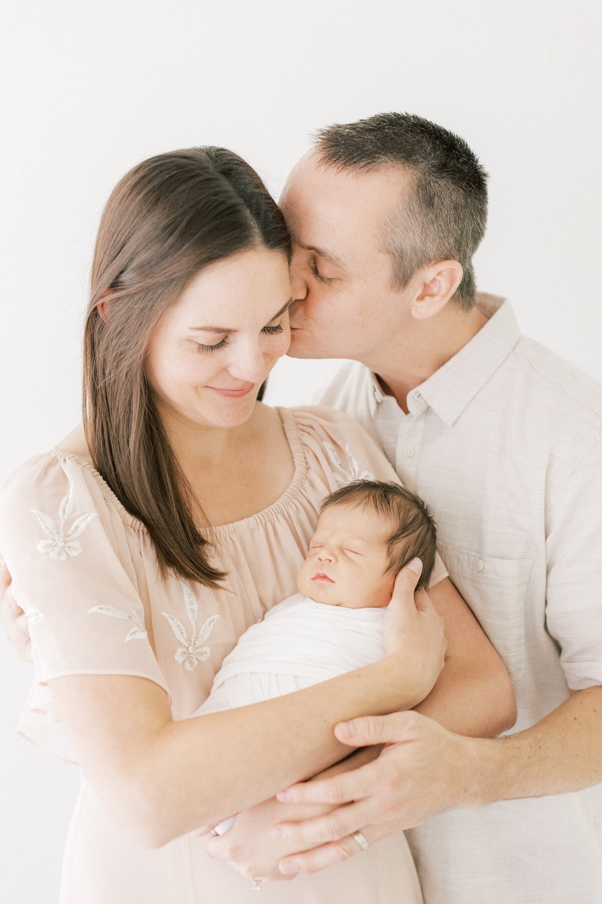 dad kissing mom on the cheek while mom holds swaddles newborn baby boy