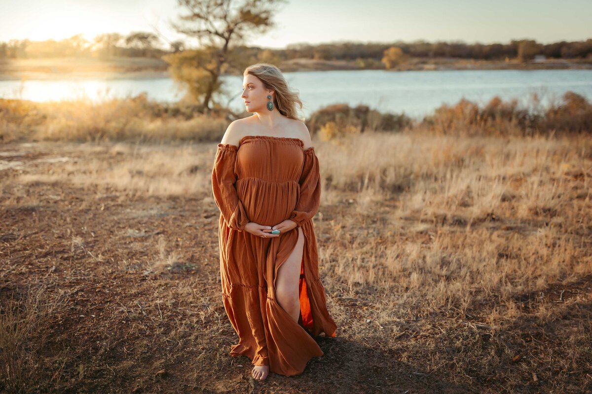 Pregnant woman standing in a field by the lake wearing a rust color dress with both her hands under her belly